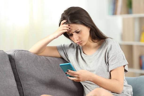 Sad teen reading bad news in a smart phone sitting on a couch in the living room at home